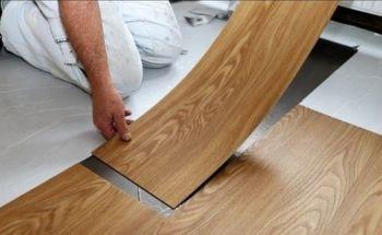  Primer plano de un instalador colocando una tabla de piso vinílico SPC de efecto madera clara, mostrando el sistema de unión click durante el proceso de instalación.