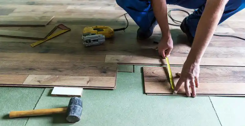 Instalación de Piso Vinílico SPC por Profesional Un instalador de pisos midiendo y colocando tablas de piso vinílico SPC de efecto madera sobre una capa de subsuelo verde, con herramientas como un martillo y una sierra.
