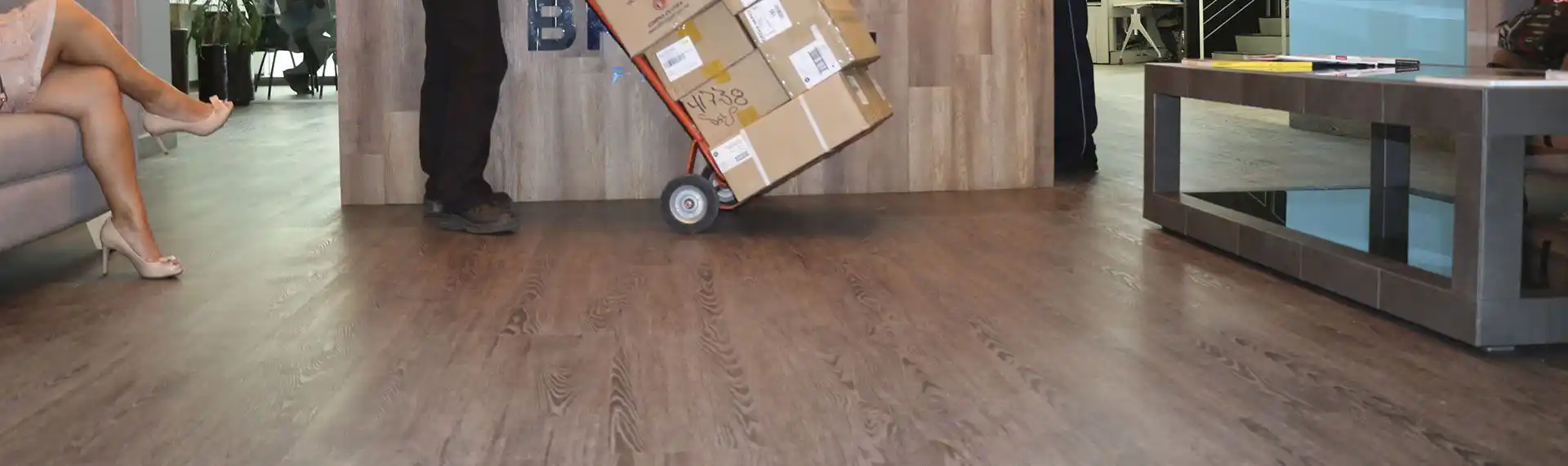 Piso vinílico SPC de efecto madera en una oficina moderna, mostrando una persona empujando un carrito con cajas, lo que demuestra la resistencia del piso al tráfico y al desgaste.