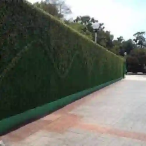 Stand de exposición de follaje artificial en una feria comercial, mostrando diferentes tipos de follaje verde y rojo, esferas decorativas y enredaderas, con personal de la empresa vistiendo uniformes verdes.