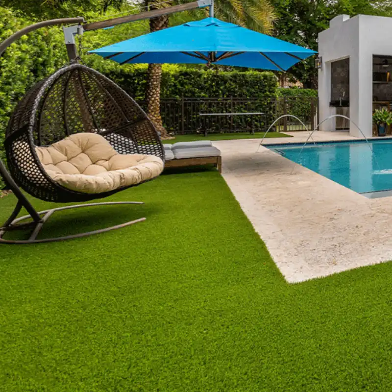 Instalación de pasto sintético modelo Luxury Elite de Grupo Orvi alrededor de una piscina en una residencia en Lomas de Angelópolis. Se aprecia un columpio de huevo, una sombrilla azul y la superficie segura y de alto drenaje ideal para zonas de alberca e infantiles.