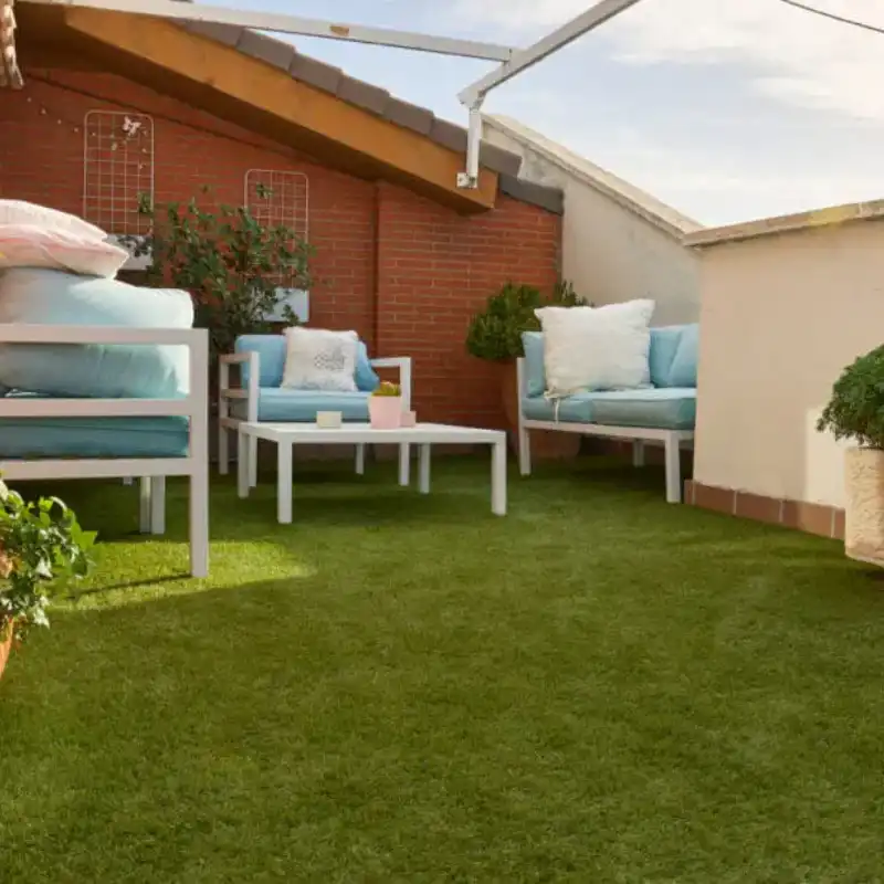 Terraza de penthouse en La Vista Country Club con pasto sintético instalado sobre losa, destacando un área de descanso con muebles de exterior blancos y cojines azules. El pasto resiste sol y viento, ideal para roof gardens y soláriums.